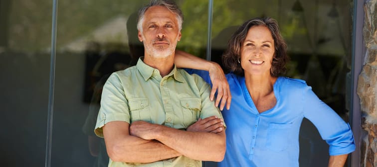 Older-aged couple standing by a sliding glass door with confident wide smiles.