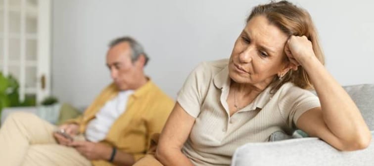 Woman looking disappointed and bothered on couch while man using smartphone in background.