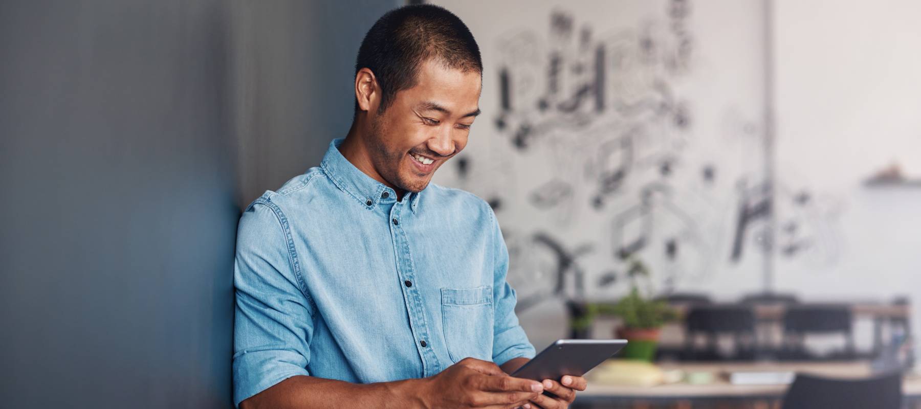 Casually dressed young Asian designer smiling and using a digital tablet while leaning against a gray wall in a modern office